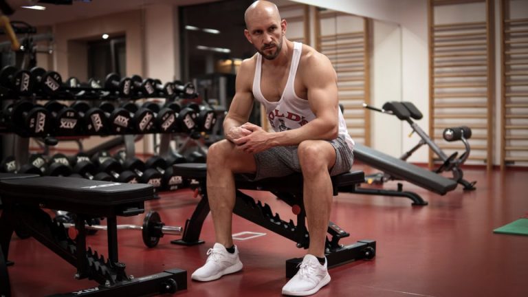 Strong, determined man in white tank top sitting in gym, showcasing fitness and strength.